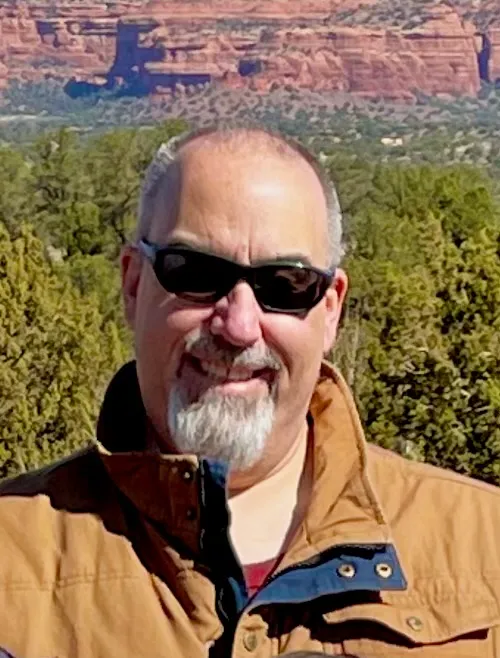 Portrait of a smiling man wearing sunglasses and a tan jacket, standing outdoors with a scenic backdrop of red rock formations, lush green trees, and clear blue skies.